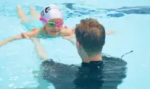 Young girl in goggles swimming toward an instructor in a pool lesson.